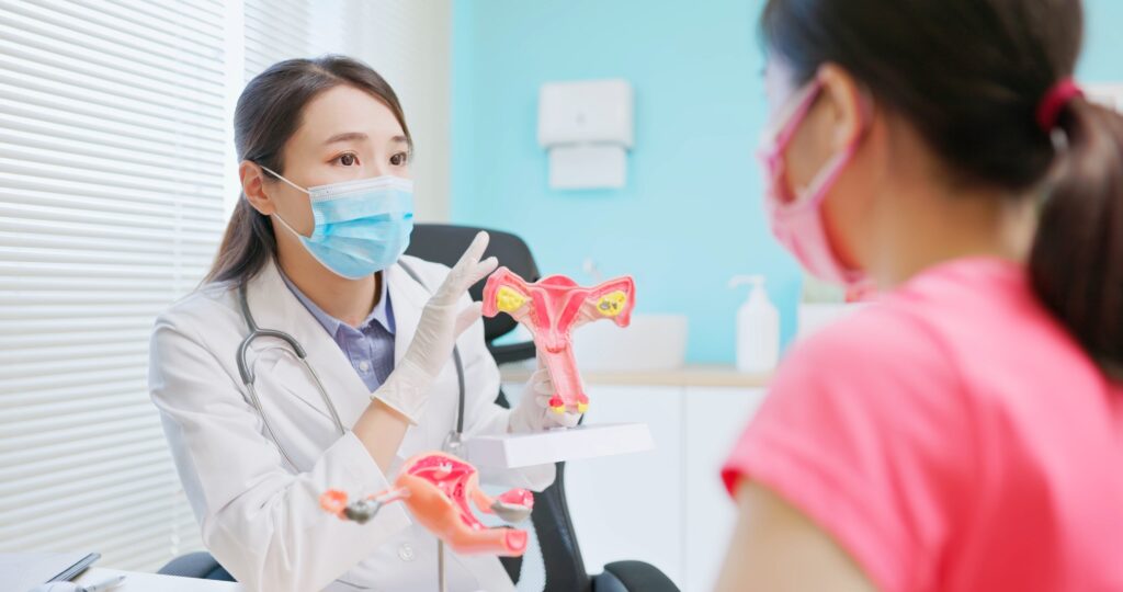 A masked female doctor discusses hormonal health with a patient, using an anatomical model of the female reproductive system in her office.