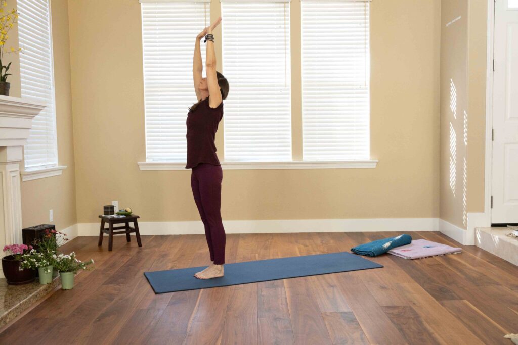 Woman standing in Mountain Pose (Tadasana), a grounding yoga pose, raising her arms upward in a peaceful indoor yoga setting to relieve lower-back pain