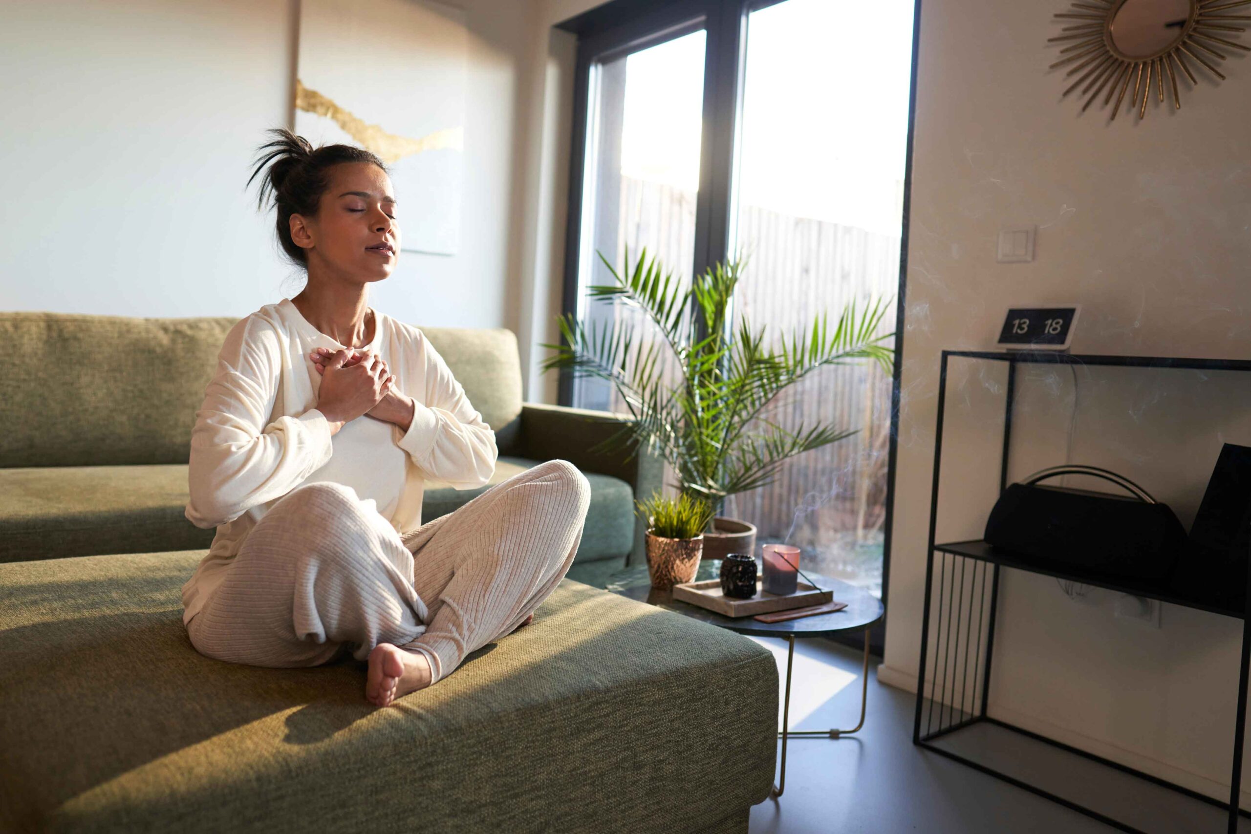 Woman practicing stress-relieving yoga at home, sitting cross-legged on a sofa with eyes closed, hands placed gently over her heart, embracing mindfulness and tranquility.