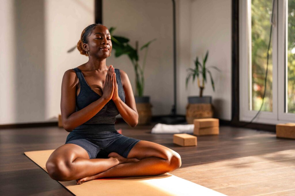 Peaceful woman practicing seated meditation yoga pose indoors, enhancing her mental health through mindful breathing and relaxation in a calming, plant-filled environment.