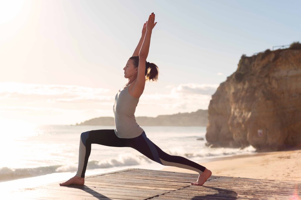 Woman practicing heart-opening yoga on a sunlit beach boardwalk, holding Warrior I with arms lifted and chest expanded toward the sky.